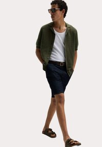 Camisa de manga corta a rayas verdes sobre una camiseta blanca, combinada con pantalones cortos de mezclilla oscura y sandalias marrones. El modelo posa con las manos en los bolsillos.