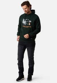 Felpa verde scuro con una stampa grafica di alberi e un caminetto; abbinata a pantaloni cargo neri con molteplici tasche con zip.