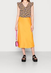 Blouse à imprimé floral avec manches à volants, assortie à une jupe midi en satin orange. Sandales compensées noires et un petit sac à main rose complètent la tenue.