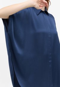 Marineblå kortærmet bluse lavet af glat stof, med krave og sideskår for en afslappet pasform. Minimalistisk design med rene linjer.