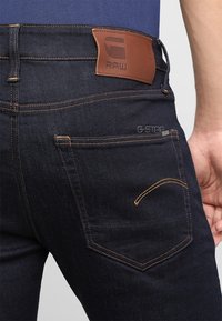 Jeans de mezclilla oscura con costuras de cobre, que presentan un parche de cuero marrón claro con logo en la cintura y detalles en el bolsillo trasero.