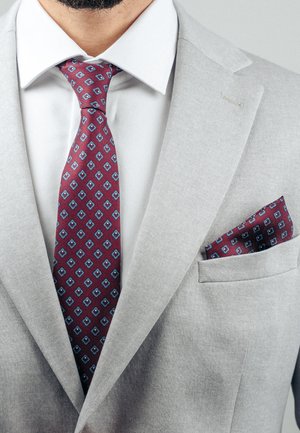 Mees, kes kannab helehallist ülikonnajakki, valget särki ja burgundiapunast lipsu sinise rombikujulise mustriga, sobiva taskurätikuga jakk-taskus.