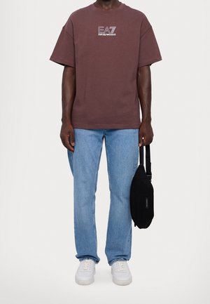 Hnědé bavlněné tričko s bílým logem, kombinované se světle modrými džínami a bílými teniskami. V pravé ruce černá taška.