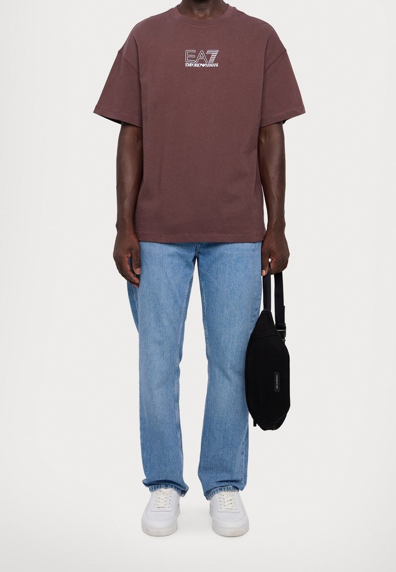 Brązowa bawełniana koszulka z białym logo, zestawiona z jasnoniebieskimi dżinsami i białymi trampkami. Czarna torba trzymana w prawej ręce.