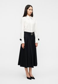 Λευκό πουκάμισο με γιακά και μακριά μανίκια, σε συνδυασμό με μαύρη φούστα σε σχήμα Α, με φαρδιά ζώνη και φιόγκους στα μανίκια. Μαύρα μυτερά παπούτσια.