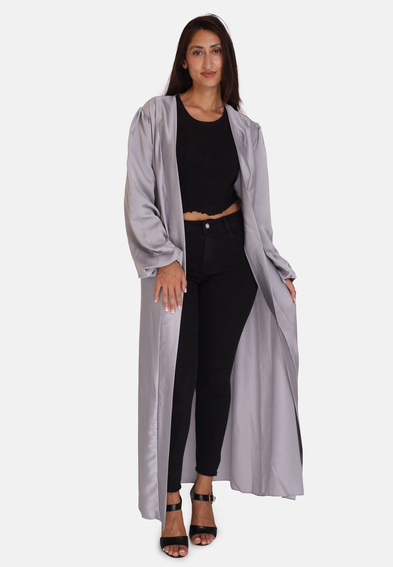 Lungo accappatoio grigio in satin con maniche lunghe, apertura frontale, caratterizzato da una consistenza liscia, abbinato a un top nero corto e pantaloni neri. Sandali neri con tacco.