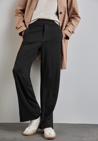Pantalons larges noirs en matériau doux, dotés de détails de couture avant et d'une taille élastique, associés à des baskets blanches et beiges.