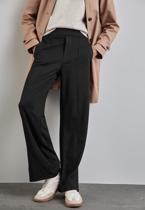 Black wide-leg pants made from a soft material, featuring front seam details and an elastic waistband, paired with white and beige sneakers.