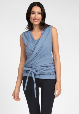 Kvinde med mørkt hår iført en ærmeløs blå omslagstop bundet i taljen og sorte leggings, smilende mod en ensfarvet baggrund.