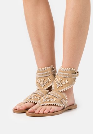 Sandali in suede beige con ricami floreali decorativi, accenti di perline, punta aperta e cinghie alla caviglia con fibbia regolabile. Suola piatta.