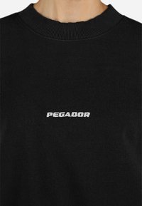Čierny sveter s rebrovaným výstrihom; na prednej strane má vyšívané biele logo s nápisom "PEGADOR". Mäkká textura a štandardný strih.