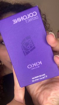 Person hält eine lila Kiko Milano Cologne District Parfümverpackung, teilweise das Gesicht bedeckend, und zeigt das Produkt nah an der Kamera.