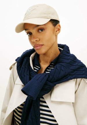 Młoda osoba nosząca beżową czapkę z daszkiem, granatowy sweter warkoczowy zarzucony na ramiona, czarno-białą pasiastą koszulę oraz lekki trencz.