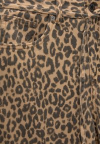 Gros plan d'un tissu avec un motif léopard marron et noir, montrant la poche avant, les coutures, le bouton en métal et les détails des rivets sur le pantalon.