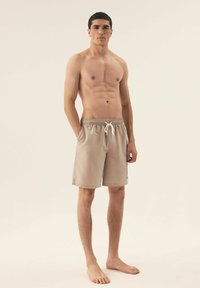 Beżowe szorty kąpielowe z siateczkową teksturą, elastycznym paskiem w talii i sznurkiem. Zawierają mały niebieski akcent z boku. Model jest bez koszulki.