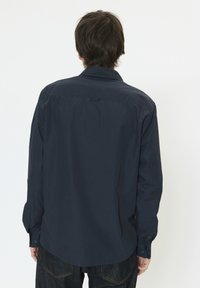 Chemise bleu marine à manches longues en tissu lisse avec un col boutonné, présentant un léger brillant et un ourlet arrondi, portée avec un denim foncé.