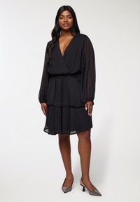 Czarna sukienka z długimi rękawami, głębokim dekoltem w kształcie litery V, projektowana w warstwy, z teksturowanej tkaniny o subtelnym wzorze kropek, zestawiona z szpilkami z akcentami srebra.