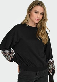 Črna majica s dolgimi rokavi z barvitimi cvetličnimi vezeninami. Mehka tkanina, sproščen kroj, rebrast ovratnik in rob.