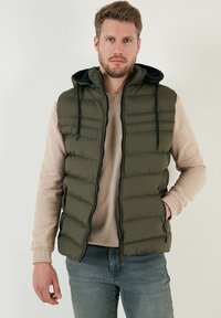 Olivgrön puffer väst med huva, framsida med dragkedja och sidofickor; bärs över en ljusbeige tröja och tillsammans med blå jeans.