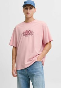 Lyserød kortærmet t-shirt lavet af bomuld, med et centralt grafisk design med ordene "Hunt Money" i maroon. Bæret med lyseblå jeans.