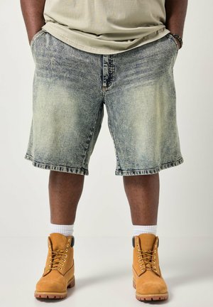Mand iført falmede denimshorts, tanfarvede arbejdstøvler med hvide sokker, beige t-shirt og armbånd, står med hænderne i lommerne.