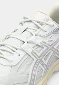 Λευκό αθλητικό παπούτσι με δίχτυ και συνθετικό επικάλυμμα, που διαθέτει ιριδίζουσες λεπτομέρειες, επίπεδα κορδόνια και υφασμένο πάτο με στρογγυλό σχήμα.