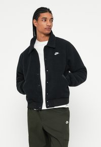Mladenič z zapletenimi lasmi nosi črno Nike flis jakno, belo majico in olivne cargo hlače, gleda levo z rokama v žepih.