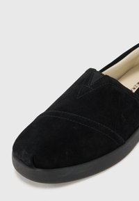 Scarpa slip-on in suede nera con punta arrotondata, caratterizzata da cuciture sottili e suola in gomma. Texture liscia e design minimale.