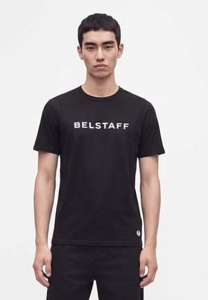 Młody mężczyzna w czarnej koszulce z napisem "BELSTAFF" na piersi, stojący na jednolitym jasnym tle.