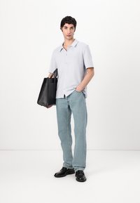 Mlad moški v svetlo sivi majici s kratkimi rokavi, svetlih modrih jeans hlačah, črnih čevljih, drži črno platneno torbo in stoji proti beli ozadju.