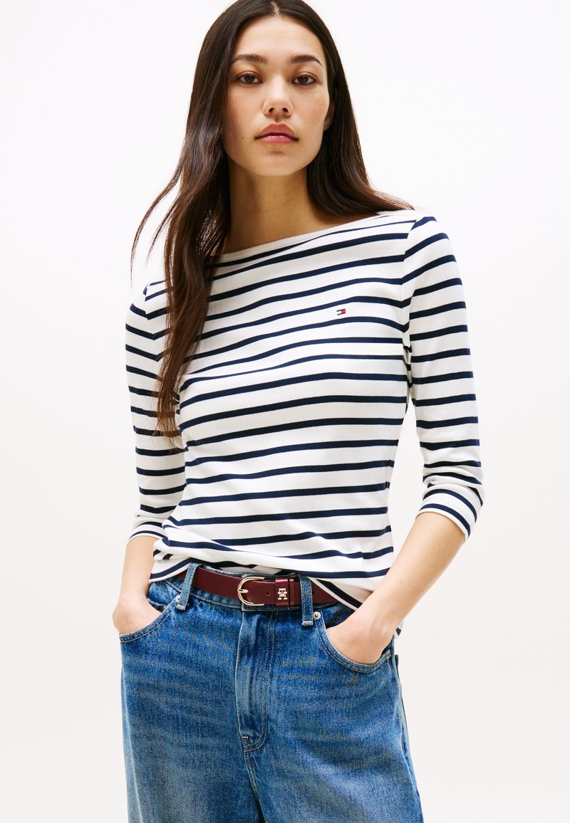 Jonge vrouw draagt een wit en navyblauw gestreept shirt met lange mouwen, blauwe spijkerbroek en een bruine riem, staat met handen in de zakken.