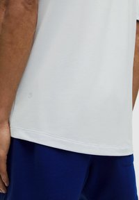 Biała koszulka z krótkimi rękawami, o gładkiej fakturze, zaokrąglonym dole i małym detalem z logo blisko dołu. Połączona z niebieskimi spodniami.