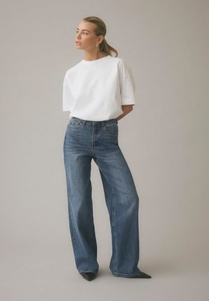 Femme aux cheveux tirés en arrière portant un t-shirt blanc, un jean bleu taille haute à jambes larges, et des chaussures noires pointues, debout devant un fond uni.