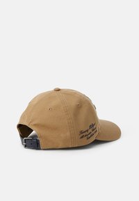 Brown cotton baseball cap with a curved visor, adjustable leather strap, and embroidered text on the side. Features a rounded top.