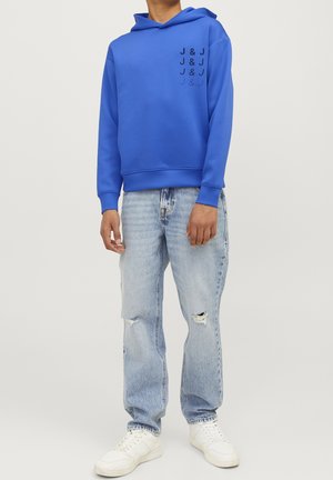 Niebieska bluza z kapturem, z pionowym wzorem czarnego tekstu z przodu. Dobranie z jasnymi, podartymi dżinsami i białymi trampkami.