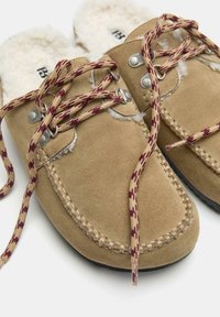 Bež suede moccasin copati z bordo in bež vzorčastimi vezalkami ter belo puhasto podlogo na belem ozadju.
