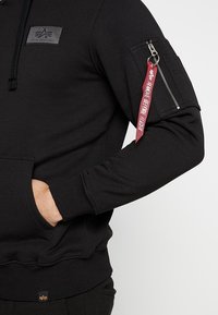 Felpa nera con cappuccio e tasca frontale, polsini a coste e tasca con zip sulla manica con etichetta in tessuto rossa. Materiale morbido e design aderente.