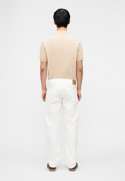 Άνδρας που φοράει μπεζ πόλο μπλουζάκι, λευκό τζιν με δερμάτινο πάτσο και σκούρα παπούτσια στέκεται με την πλάτη σε απλό φόντο.