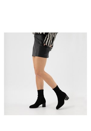 Persona che indossa stivaletti neri con tacco a blocco, una gonna in pelle nera e un maglione a righe bianche e nere, in piedi su uno sfondo bianco.