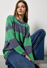 Grüner und marineblauer gestreifter Pullover mit lockerer Passform, rundem Halsausschnitt und strukturierter Strickart. Kombiniert mit hellblauen weiten Jeans.