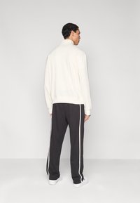 Λευκό φούτερ με ψηλό γιακά, σε συνδυασμό με σκούρο γκρι παντελόνι που έχει λευκές ρίγες στο πλάι. Το μοντέλο βρίσκεται με την πλάτη προς το μέρος της κάμερας σε απλό φόντο.