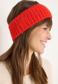 Czerwona dziana opaska na głowę o grubym, strukturalnym wzorze. Miękki materiał z prążkowanym wzorem, noszona na włosach w widoku z profilu.
