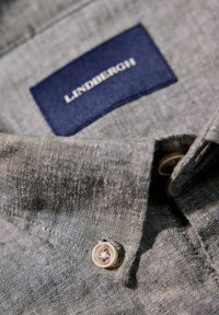 Grijze linnen overhemddetail met een marineblauwe label met "LINDENBERG." Bevat een enkele knop met een subtiel ontwerp op de kraag.