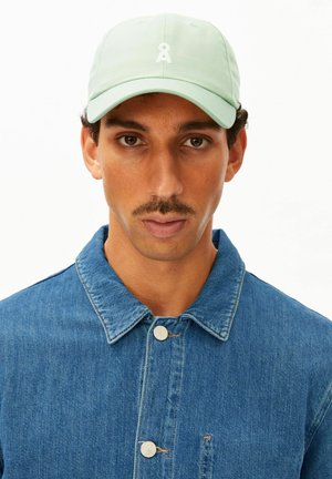 Mann mit braunen Augen und Schnurrbart trägt eine hellgrüne Kappe und eine blaue Jeansjacke vor weißem Hintergrund.
