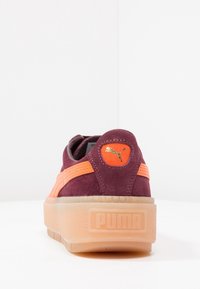 Baskets Puma en daim violet foncé, avec des accents orange vif et une semelle extérieure en gomme texturée. Présentent un logo doré sur la languette arrière.