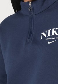 Mörkblå kvartslång dragkedja sweatshirt i mjukt material, med en Nike-logotyp och texten "Athletic dept. 1928" i vitt. Slät textur.