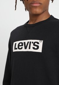 Sudadera negra con un gran logo blanco de Levi's en el pecho. La tela es suave, con un cuello acanalado y mangas largas.