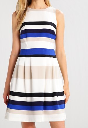 Femme portant une robe sans manches, longueur genou, à rayures horizontales noires, bleues, beige et blanches, debout devant un fond uni.