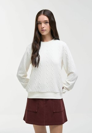 Femme aux longs cheveux foncés porte un pull en maille torsadée blanc et une jupe marron avec des poches avant, debout devant un fond blanc uni.