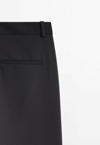 Le pantalon noir présente une texture lisse, une taille ajustée avec passants de ceinture et une poche arrière avec un détail de couture subtil.